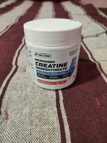 Customer's white tub of Wellcore Micronised Creatine Monohydrate, Watermelon Wave flavor, on a striped surface.