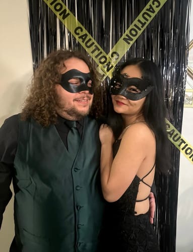 Reviewer and a guest in black masquerade masks and formal wear posing in front of a caution tape backdrop at their party.