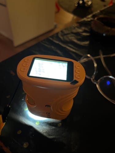 Rezensent zeigt ein leuchtendes, orangefarbenes Handgerät mit eingeschaltetem Bildschirm auf einem Tisch.