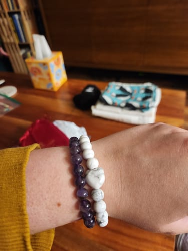 Customer's wrist wearing a white Howlite bracelet with a heart bead next to a purple beaded bracelet.