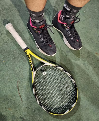 A reviewer's black Nike S.T. Flare EP Basketball Shoes with pink accents, seen from above on a green court with a tennis racquet.