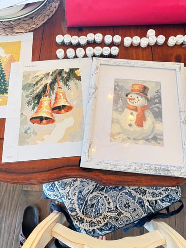 Reviewer's completed Festive Village painting of a snowman in a white frame, next to an in-progress canvas of bells.