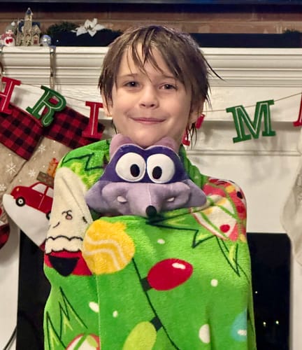 Customer's happy child wrapped in a blanket, holding the purple Original Derek Plushie in front of a fireplace.