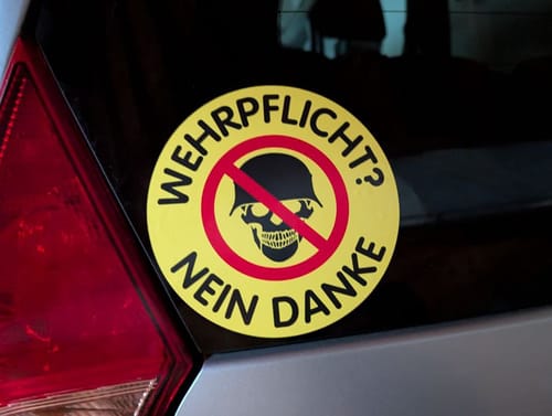 Rezensent zeigt den runden „Wehrpflicht? Nein Danke“-Aufkleber auf einer Autoscheibe.