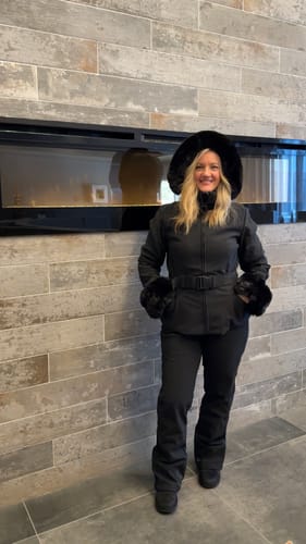Customer smiling in the black faux fur ski jacket, showing its fitted, belted style indoors.