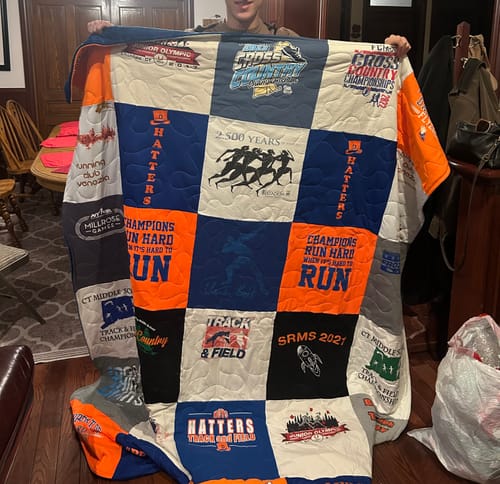 A person holding up a large Premium T-Shirt Quilt made from colorful blue, orange, and white track and field shirts.