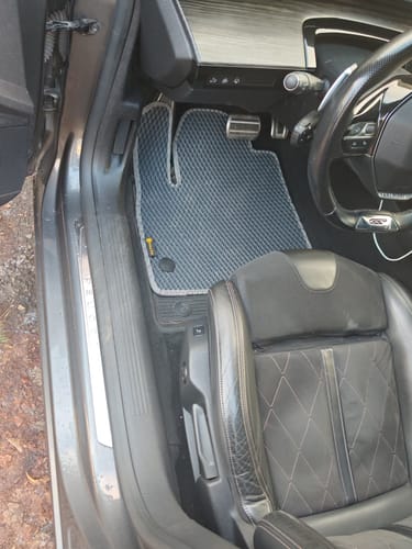 Klient pokazuje szary, dopasowany dywanik samochodowy zamontowany po stronie kierowcy w ciemnym wnętrzu auta.