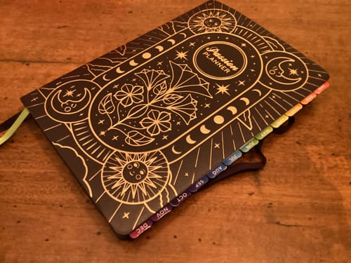 Customer's colorful Monthly Tabs attached to the side of a black celestial-themed Passion Planner.