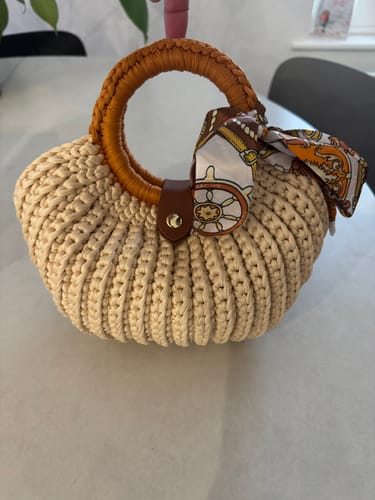 Customer's completed beige Shell crochet Handbag with a round wrapped handle and a decorative scarf tied on, resting on a table.