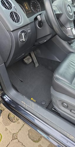 Recenzent pokazuje idealnie dopasowany czarny dywanik samochodowy po stronie kierowcy.