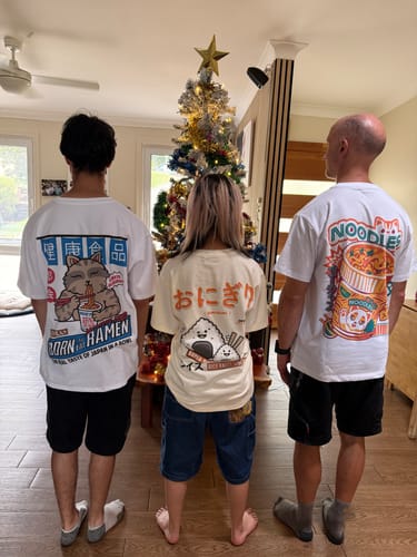 Three customers showing the back prints of their Neko Noodles T-Shirts in front of a Christmas tree.