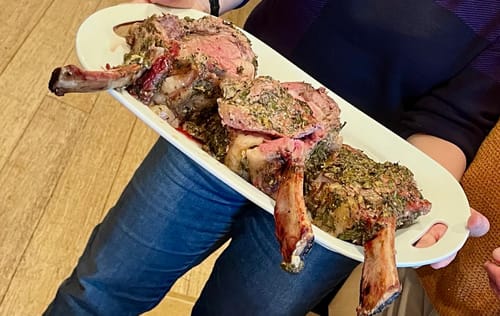 Customer holding a white platter with a cooked, herb-crusted Tomahawk Prime Rib Roast, sliced for serving.