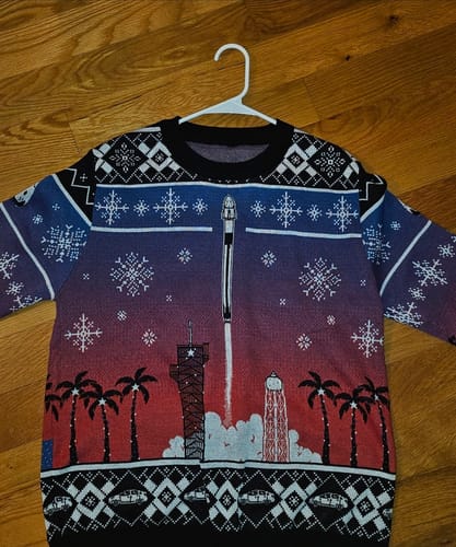 Customer's Crew Launch Christmas Sweater on a hanger, showing its detailed space-themed holiday design with a rocket launch.