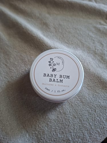 Customer's round tin of Baby Bum Balm resting on a light brown fabric.