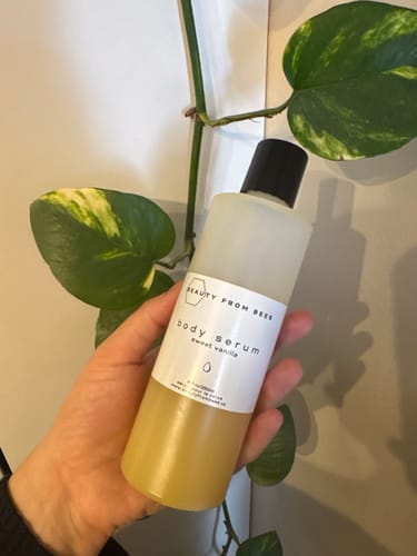 Customer's hand holding the Sweet Vanilla Body Serum bottle against a leafy plant background.