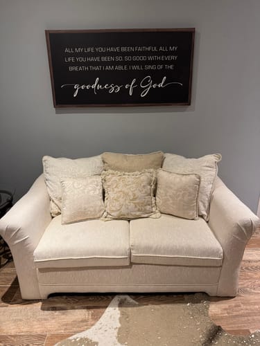 Reviewer's 'Goodness Of God Faith Sign II' canvas hanging on a gray office wall above a cream-colored loveseat.
