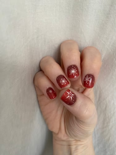 Hand einer Kundin mit kurzen Nägeln, die das rote Ombré-Design des UV Gels Winter Rouge mit Schneeflocken zeigen.