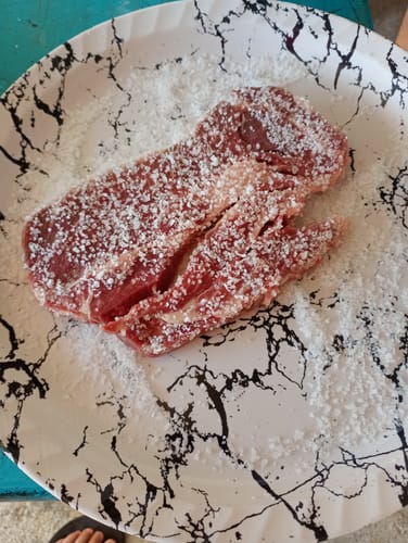 Cliente preparando un bistec de diezmillo crudo, cubierto con sal en un plato marmoleado.