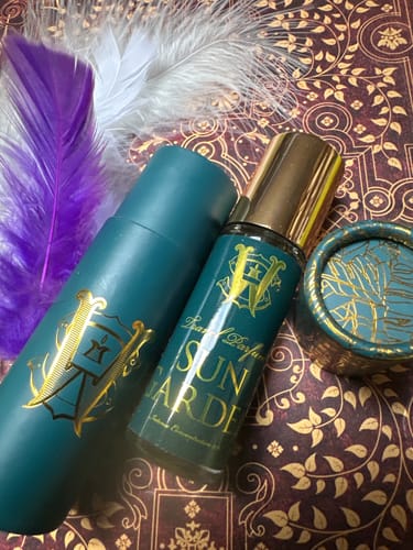 Reviewer's Sun Garden perfume bottle and its teal packaging tube, shown on an ornate background with two feathers.