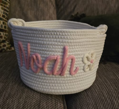Customer's personalized white basket with the name 'Noah' in pink and a small flower sitting on a couch.