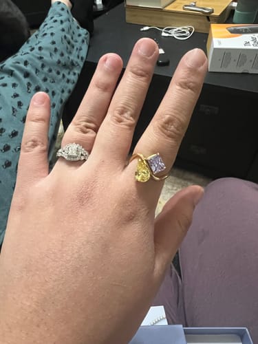 A person's hand wearing the Navae Birthstone Ring with a yellow pear-shaped and a light purple square-cut stone.