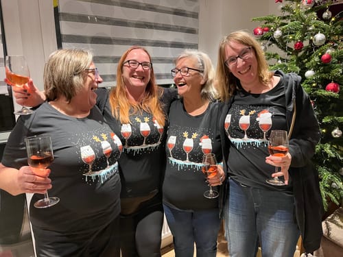 Vier Kundinnen tragen die passenden schwarzen „Weihnachtswein“-Shirts und stoßen neben einem Weihnachtsbaum mit Wein an.