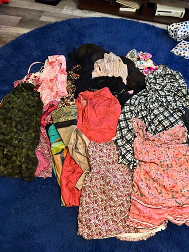 Reviewer's large assortment of wholesale clothing from their order, including flannel shirts and dresses, on a rug.