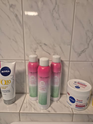 Rezensent zeigt drei Flaschen des Haarentfernungsschaums auf einer gefliesten Ablage im Badezimmer.