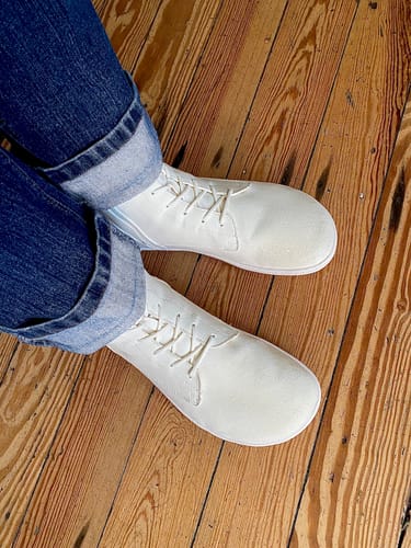 Draufsicht eines Rezensenten, der die cremefarbenen Ambition II Winter Barfußschuhe mit Jeans auf einem Holzboden trägt.