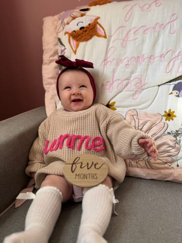 Customer's baby smiling in a beige Hand Knitted Personalized Sweater with a name embroidered in pink.