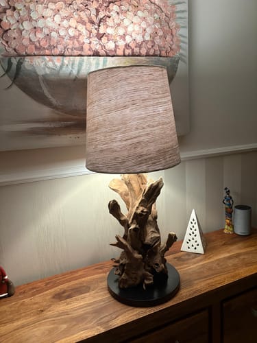 Rezensent zeigt die rustikale Wurzelholz Tischlampe IKAL auf einem Holztisch. Die Lampe ist eingeschaltet und spendet warmes Licht.