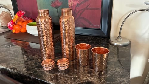 Reviewer's two hammered Himalayan Ayurvedic Pure Copper Bottles and two matching cups on a marble countertop.