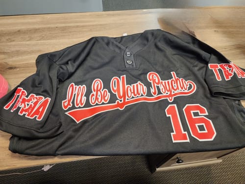 Customer's custom black and red softball jersey with "I'll Be Your Psycho 16" on the front, laid on a wooden desk.