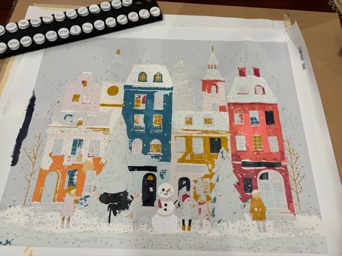 Customer's in-progress Snowy Games Paint by Number canvas showing a colorful, snowy village scene with paints nearby.