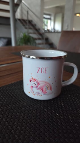 Rezensent zeigt die personalisierte Einhorn-Emaille-Tasse mit dem Namen 'Zoe' auf einem dunklen Tischset.