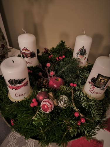 Rezensent zeigt die vier Adventskerzen mit Weihnachtsmotiven in einem geschmückten, grünen Adventskranz.