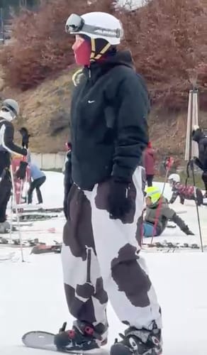 Reviewer standing on a snowy slope wearing the baggy cow print snow pants with a snowboard.