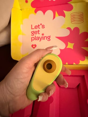 Reviewer holding the green, avocado-shaped Avo Clitoral Massager in their hand in front of its colorful packaging.