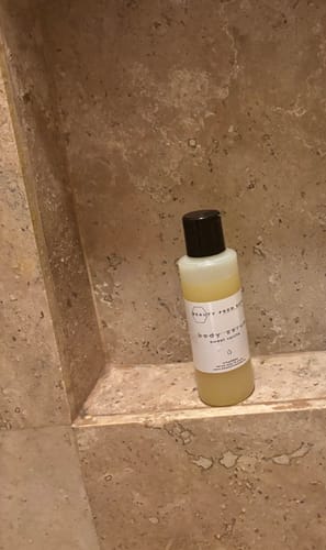 Reviewer's travel-size bottle of Body Serum sitting on a tiled shower shelf.
