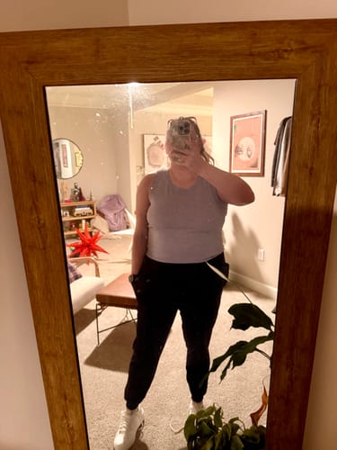 Reviewer takes a mirror selfie wearing the mystery item, a light purple tank top, paired with black pants.
