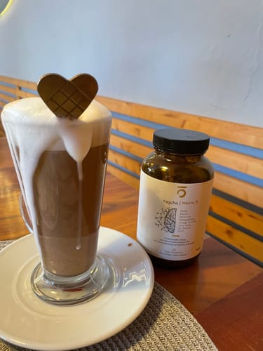 Eine Kundin zeigt das Glas des Anti-Stress Komplex neben einem fertigen, schaumigen Kakaogetränk auf einem Holztisch.