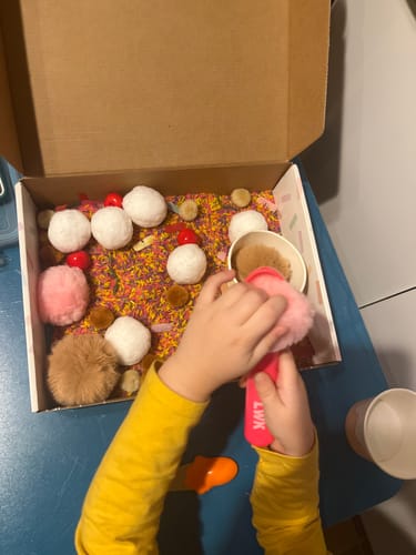 A customer's child plays with The Sensory Box, scooping colorful rice and pom-poms with a pink tool.