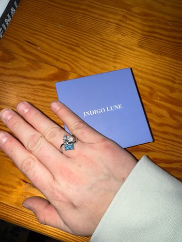 A reviewer's hand wearing the Navae Birthstone Ring, featuring a clear pear-cut stone and a light blue radiant-cut stone.