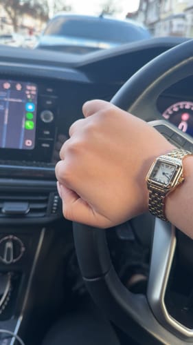 A customer wearing a classy gold watch with a square face while holding a steering wheel.