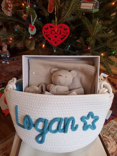Customer's white knitted basket, personalized with 'Logan' and a star icon in blue yarn, shown filled with gifts.