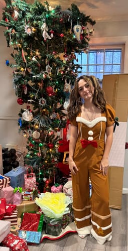 A reviewer shows the gingerbread jumpsuit's fit on a person standing in a festive setting by a Christmas tree.