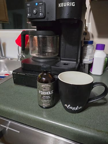 Customer's bottle of Foxhole Coffee Concentrate on a kitchen counter next to a black mug and a Keurig coffee maker.