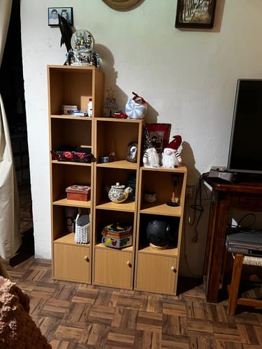 Un cliente muestra el juego de 3 estanterías modulares de color madera claro, montadas en forma de escalera en una habitación y llenas de objetos decorativos.