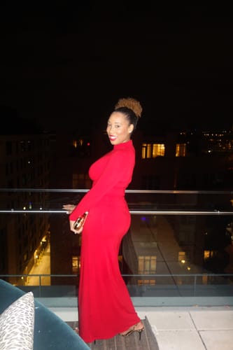 Customer posing sideways in the red NW Turtleneck Draped Tie Dress on a balcony at night.