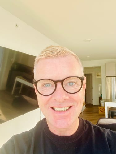 A close-up selfie of a smiling reviewer wearing glasses indoors.